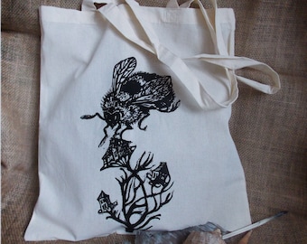 Op de afbeelding: Een witte tote bag met een zwarte print van een hommel die boven een plant met kleine huisjes zweeft. De hommel is groot en gedetailleerd, met zichtbare vleugels en poten. De plant is gestileerd en heeft een dikke stam met takken.