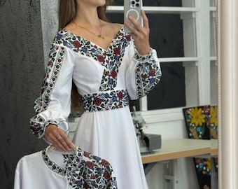 May include: White dress with floral embroidery on the sleeves, bodice, and waist. The dress has long sleeves and a flowing skirt. A matching white blouse with similar embroidery is held in front of the dress.