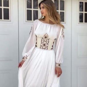 May include: A floor-length white dress featuring long, loose sleeves with embroidered cuffs and shoulders. The garment includes a structured, cream-colored corset-style bodice adorned with intricate brown and black geometric embroidery patterns.