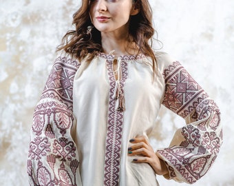 May include: A white embroidered blouse with long sleeves and a pink floral pattern. The blouse has a round neckline and a tie at the neck.