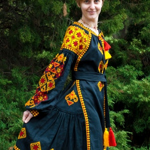 Op de afbeelding: Een zwarte jurk met lange, volumineuze mouwen en een geplooide rok, versierd met ingewikkelde gele en rode geometrische borduurpatronen. Het kledingstuk is voorzien van een bijpassende strikceintuur en decoratieve rode en gele kwastjes bij de hals.