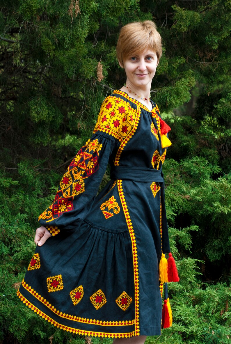 Op de afbeelding: Een zwarte jurk met lange, volumineuze mouwen en een geplooide rok, versierd met ingewikkelde gele en rode geometrische borduurpatronen. Het kledingstuk is voorzien van een bijpassende strikceintuur en decoratieve rode en gele kwastjes bij de hals.