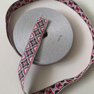 Op de afbeelding: Een rol grijs lint met een decoratief geweven patroon in rood, zwart en wit. Het lint is afgerold en toont het gedetailleerde ontwerp van de rand. Het lint wordt waarschijnlijk gebruikt voor knutselen, naaien of het verfraaien van diverse projecten.