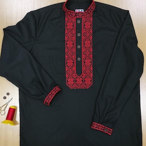 May include: Black long-sleeve shirt with red embroidered floral patterns on the collar, front, and cuffs. The shirt has a mandarin collar and buttoned cuffs. Sewing supplies, including scissors, thread, and buttons, are visible.