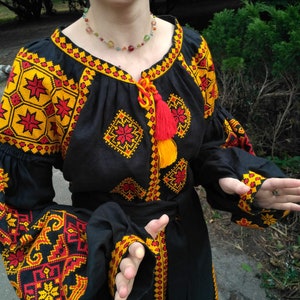Op de afbeelding: Een zwarte blouse met lange mouwen en ingewikkeld rood en geel geometrisch borduurwerk op de mouwen en borst. Het kledingstuk heeft een ronde halslijn met een striksluiting, rode en gele kwastjes en een bijpassende stoffen ceintuur.