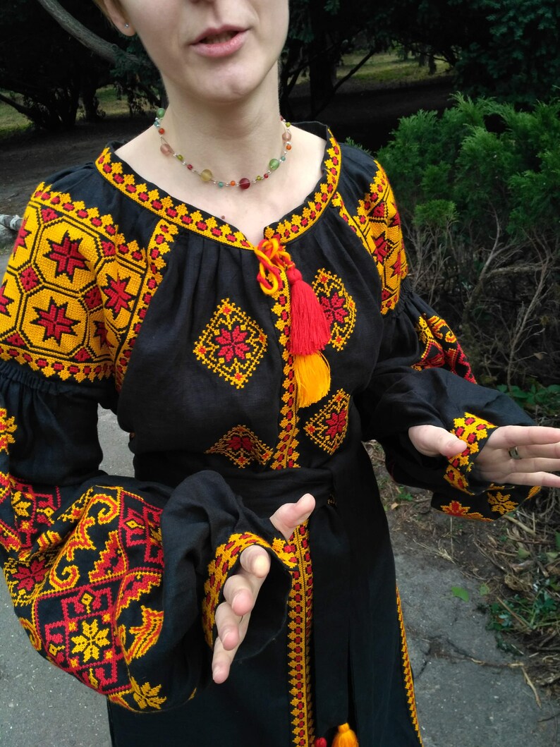 Op de afbeelding: Een zwarte blouse met lange mouwen en ingewikkeld rood en geel geometrisch borduurwerk op de mouwen en borst. Het kledingstuk heeft een ronde halslijn met een striksluiting, rode en gele kwastjes en een bijpassende stoffen ceintuur.