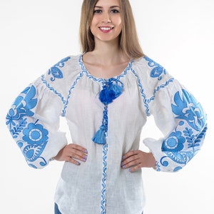 Pode incluir: Uma blusa de linho branca com padrões florais bordados em azul e um laço de borla azul. A blusa tem mangas compridas e um decote redondo.