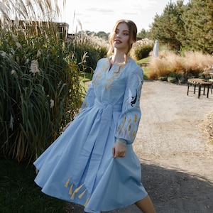 May include: A woman wearing a light blue embroidered dress with gold accents and white birds. She is standing in a grassy area with tall, dried grasses in the background.