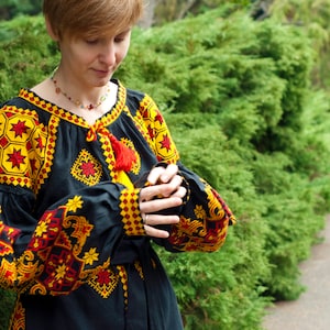 Op de afbeelding: Een zwarte blouse met lange mouwen en ingewikkeld geel en rood geometrisch borduurwerk op de schouders, borst en manchetten. Het kledingstuk heeft een ronde halslijn met een rood trekkoord met kwastjes en wijde, geplooide mouwen.