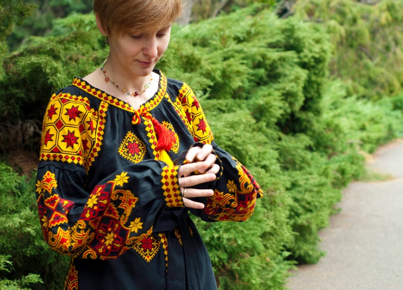 Op de afbeelding: Een zwarte blouse met lange mouwen en ingewikkeld geel en rood geometrisch borduurwerk op de schouders, borst en manchetten. Het kledingstuk heeft een ronde halslijn met een rood trekkoord met kwastjes en wijde, geplooide mouwen.