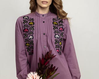 Puede incluir: Una blusa de manga larga de color morado con cuello alto y bordados florales. La blusa tiene un cierre de botones en la parte delantera y un ajuste holgado.