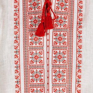 May include: A white linen shirt featuring intricate red and black geometric embroidery along the front placket. The neckline includes a split opening with red braided ties that end in two small red tassels, creating a detailed pattern.