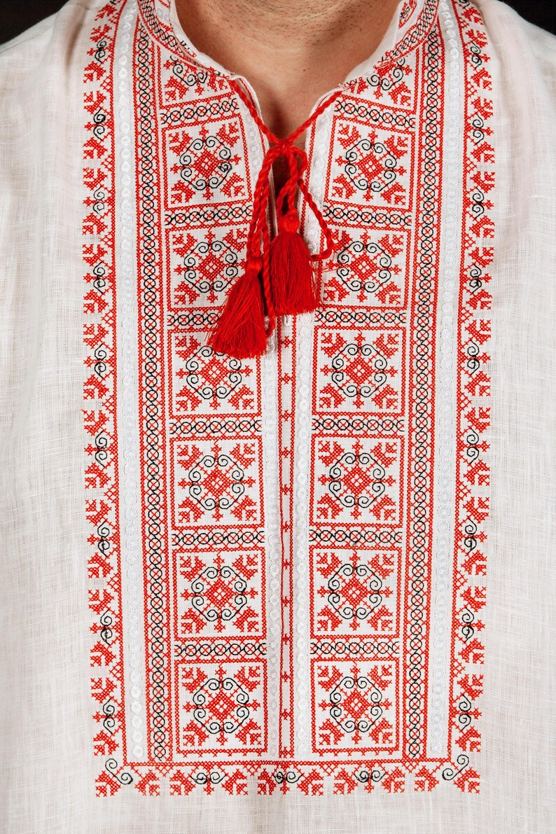 May include: A white linen shirt featuring intricate red and black geometric embroidery along the front placket. The neckline includes a split opening with red braided ties that end in two small red tassels, creating a detailed pattern.