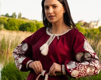 Könnte beinhalten: Eine weinrote Tunika mit weißen Blumenstickereien an den Ärmeln und am Ausschnitt. Die Tunika hat eine cremefarbene Spitzenborte und einen passenden Gürtel. Die Ärmel sind lang und die Tunika wird mit einer weißen Hose kombiniert.