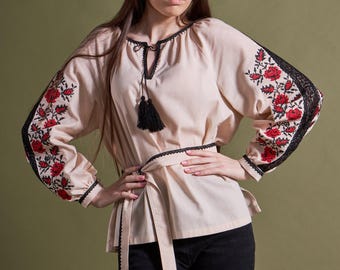 May include: A beige blouse with long sleeves featuring red floral embroidery on the sleeves. The blouse has black lace trim, a tie-waist, and a black tassel at the neckline. The model is wearing black pants.