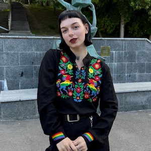 May include: A black long-sleeved shirt featuring a vibrant, multi-colored floral embroidery pattern on the chest, including two rainbow-colored unicorns. The cuffs of the sleeves are accented with horizontal rainbow stripes, and the shirt has a mandarin collar.