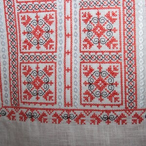 May include: A white linen fabric featuring intricate cross-stitch embroidery in vibrant red and black thread. The design consists of repeating geometric square motifs, floral-like patterns, and vertical bands of circular and linear stitching.