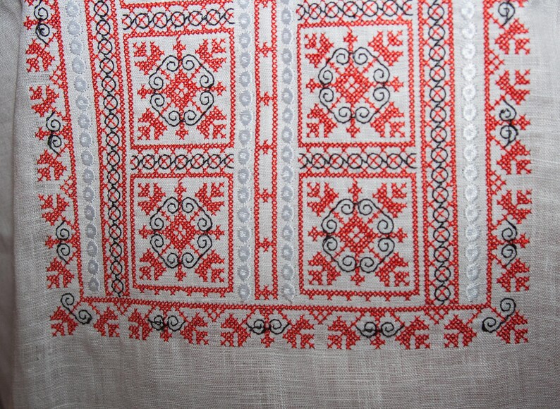 May include: A white linen fabric featuring intricate cross-stitch embroidery in vibrant red and black thread. The design consists of repeating geometric square motifs, floral-like patterns, and vertical bands of circular and linear stitching.