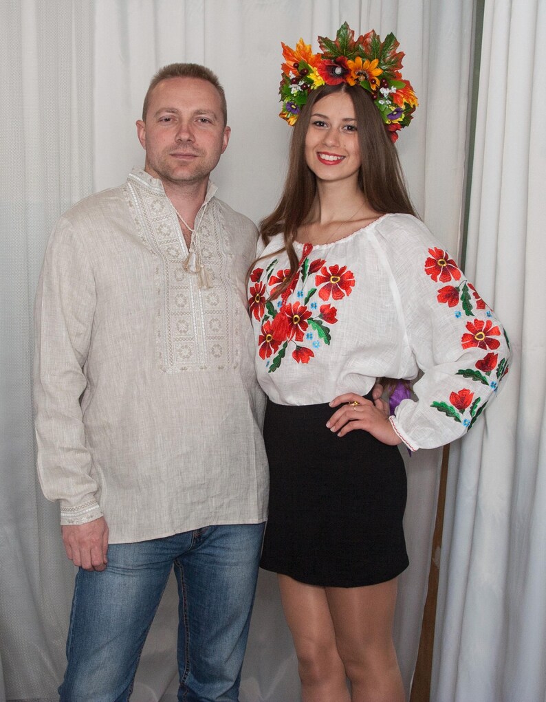 May include: A beige long-sleeved shirt with white embroidery and a white long-sleeved shirt with red and green floral embroidery, paired with a black skirt and a colorful floral crown featuring autumn-toned leaves and flowers.