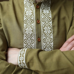 May include: Olive green long-sleeved shirt with intricate white embroidered detailing. The embroidery adorns the collar, placket, and cuffs, creating a striking contrast. The shirt features wooden buttons and a mandarin collar, offering a unique style.