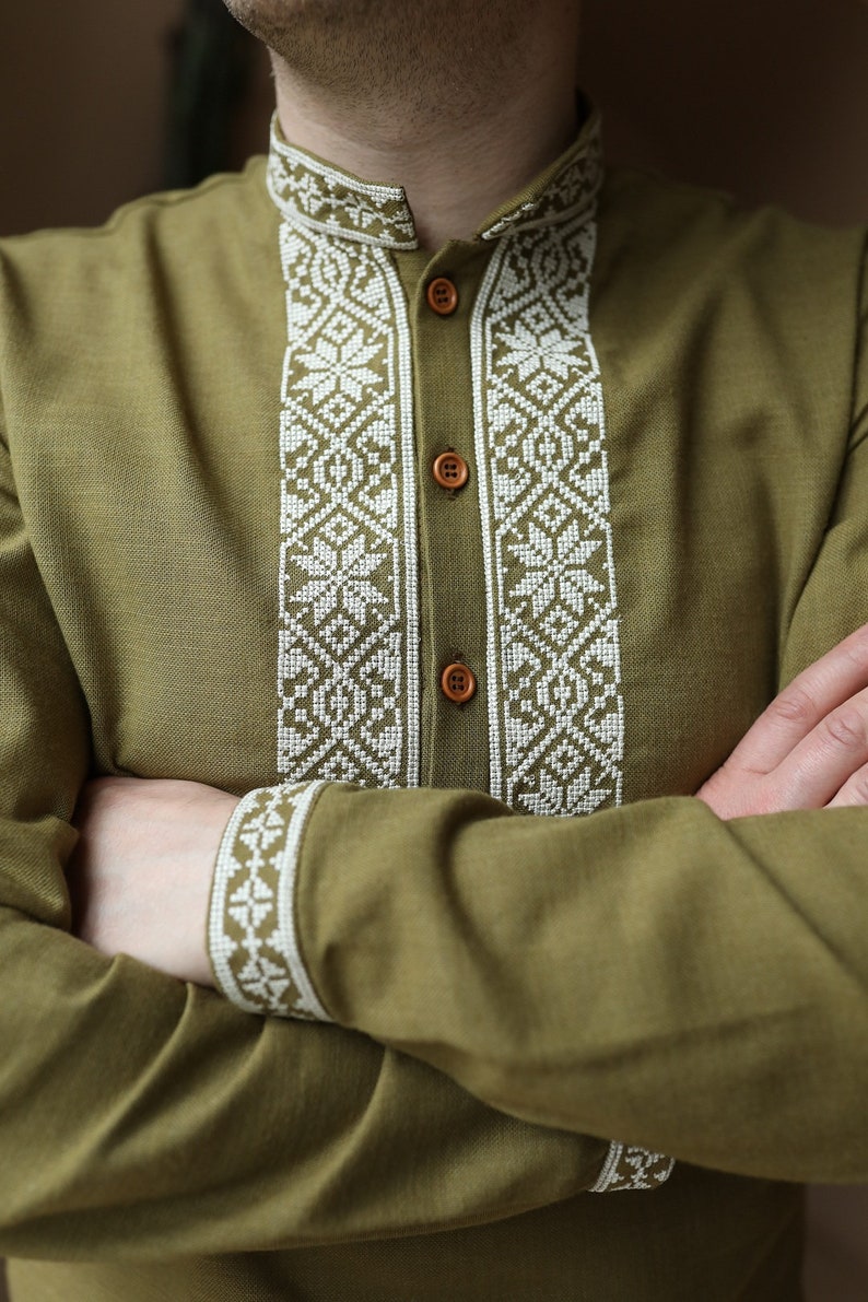 May include: Olive green long-sleeved shirt with intricate white embroidered detailing. The embroidery adorns the collar, placket, and cuffs, creating a striking contrast. The shirt features wooden buttons and a mandarin collar, offering a unique style.