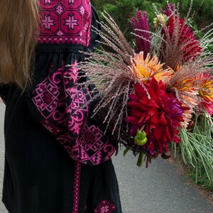 Op de afbeelding: Een zwarte tuniek met lange mouwen en felroze geometrisch borduurwerk op de schouders en mouwen, gecombineerd met een groot, kleurrijk boeket van donkerrode, oranje en paarse bloemen met lang, dun en fijn groen blad.