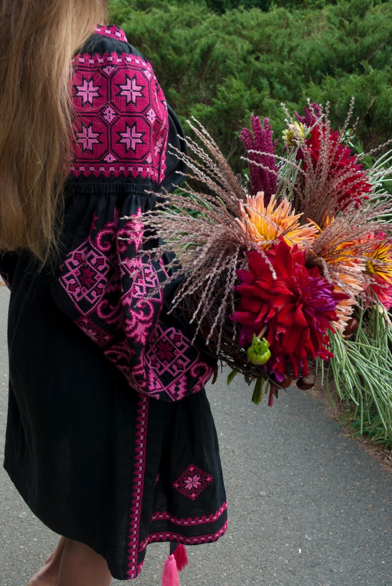 Op de afbeelding: Een zwarte tuniek met lange mouwen en felroze geometrisch borduurwerk op de schouders en mouwen, gecombineerd met een groot, kleurrijk boeket van donkerrode, oranje en paarse bloemen met lang, dun en fijn groen blad.