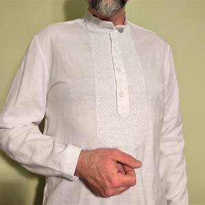 May include: White long-sleeve shirt with a mandarin collar and a decorative embroidered panel down the front. The shirt has three buttons and embroidered cuffs. The shirt is made of a lightweight fabric.