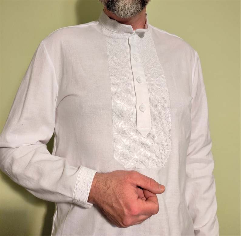 May include: White long-sleeve shirt with a mandarin collar and a decorative embroidered panel down the front. The shirt has three buttons and embroidered cuffs. The shirt is made of a lightweight fabric.