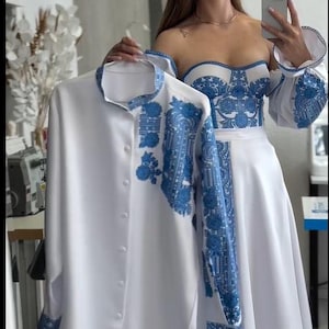 Op de afbeelding: Witte jurk en shirt set met blauwe bloemenborduurwerk. De jurk heeft een strapless lijfje en een zwierige rok. Het shirt heeft een opstaande kraag en een knoopsluiting. Beide kledingstukken hebben blauwe bloempatronen.