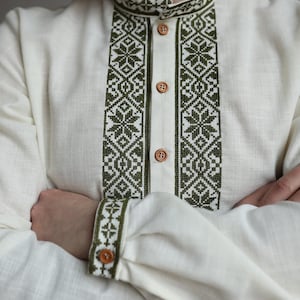 May include: Off-white linen shirt with olive green embroidered details. The shirt features a mandarin collar, button-down front, and intricate geometric patterns on the collar, placket, and cuffs. The long sleeves are cuffed with a single button.
