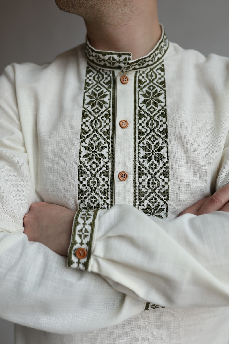 May include: Off-white linen shirt with olive green embroidered details. The shirt features a mandarin collar, button-down front, and intricate geometric patterns on the collar, placket, and cuffs. The long sleeves are cuffed with a single button.