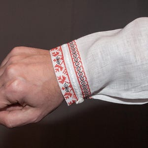 May include: A white long-sleeved shirt featuring a decorative cuff with intricate red and black geometric embroidery. The fabric has a visible linen-like texture, and the cuff is secured with a small white button, showcasing detailed needlework.
