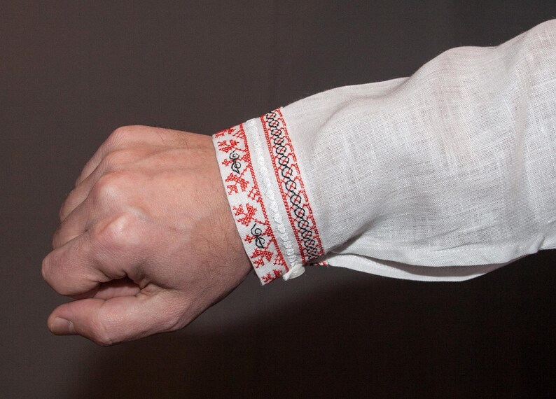 May include: A white long-sleeved shirt featuring a decorative cuff with intricate red and black geometric embroidery. The fabric has a visible linen-like texture, and the cuff is secured with a small white button, showcasing detailed needlework.