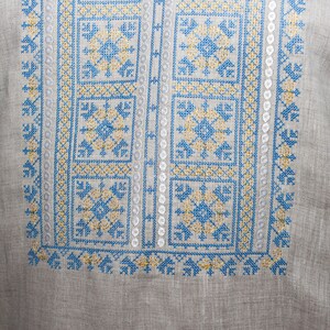 May include: A light gray long-sleeve shirt featuring intricate blue and yellow cross-stitch embroidery on the chest. The garment includes a neckline with braided drawstrings finished with light blue and yellow tassels on each side.