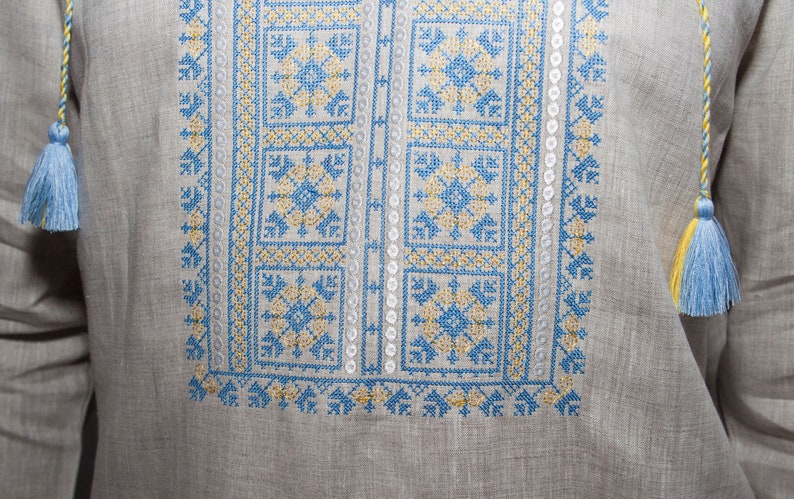 May include: A light gray long-sleeve shirt featuring intricate blue and yellow cross-stitch embroidery on the chest. The garment includes a neckline with braided drawstrings finished with light blue and yellow tassels on each side.