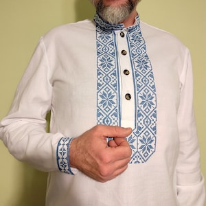 May include: White long-sleeved shirt with blue embroidered floral patterns on the collar, placket, and cuffs. The shirt has a mandarin collar and wooden buttons. The embroidery features a geometric design with snowflake-like motifs.