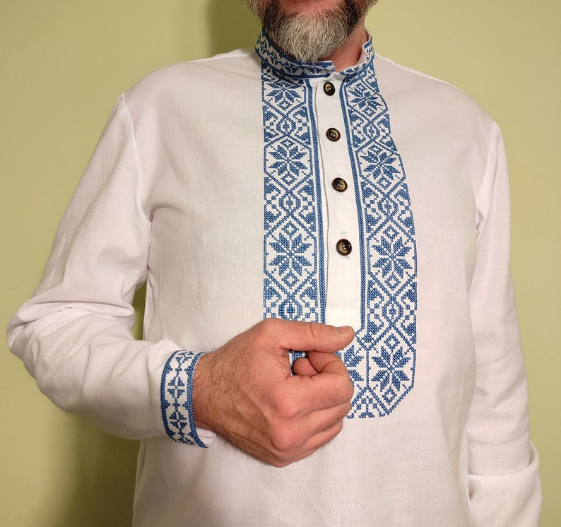 May include: White long-sleeved shirt with blue embroidered floral patterns on the collar, placket, and cuffs. The shirt has a mandarin collar and wooden buttons. The embroidery features a geometric design with snowflake-like motifs.