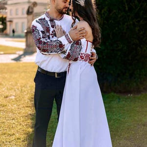 Op de afbeelding: Een wit shirt met lange mouwen en ingewikkeld rood en zwart borduurwerk op de mouwen en borst, gecombineerd met een zwarte broek en een witte vloerlange jurk met bijpassend rood en zwart borduurwerk in de taille en een gouden haaraccessoire.