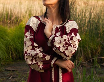 Könnte beinhalten: Burgunderfarbene Bluse mit cremefarbener Blumenstickerei auf den Ärmeln und dem Oberteil. Die Bluse hat eine Spitzenborte am Ausschnitt und an den Ärmeln sowie einen passenden Bindegürtel. Das Model trägt eine weiße Hose. Der Hintergrund ist ein Feld mit hohem Gras.