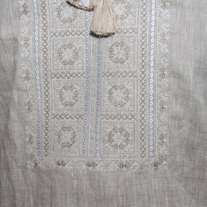 May include: A light beige linen shirt featuring a rectangular chest panel with intricate white and tan geometric embroidery. The design includes vertical columns of small circles and two beige tassels hanging from the neckline area.