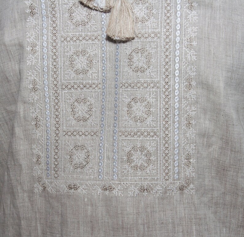 May include: A light beige linen shirt featuring a rectangular chest panel with intricate white and tan geometric embroidery. The design includes vertical columns of small circles and two beige tassels hanging from the neckline area.