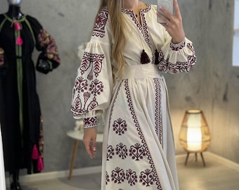 May include: A cream-colored dress with long, puffed sleeves and intricate burgundy embroidery. The dress features a V-neckline, a cinched waist, and a flowing skirt. The embroidery includes floral and geometric patterns. A woman is wearing the dress and taking a selfie.