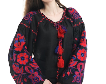 May include: A black linen blouse with red and blue embroidered floral patterns. The blouse has a V-neck and long sleeves with a tassel detail at the neckline.