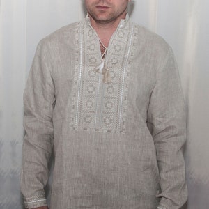 May include: A beige long-sleeved linen shirt featuring a vertical panel of intricate white and light brown geometric embroidery down the center chest. The shirt includes a mandarin collar with a drawstring closure and matching embroidered cuffs.