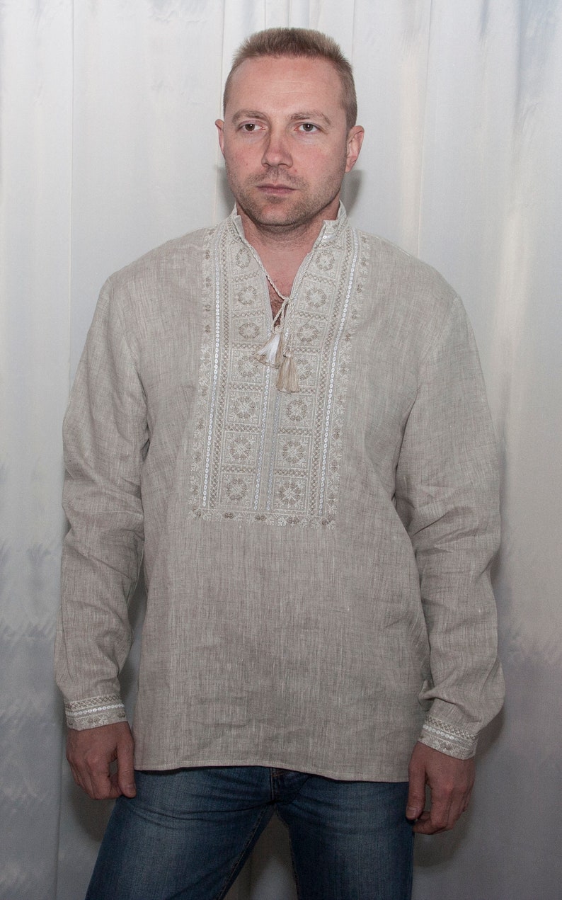 May include: A beige long-sleeved linen shirt featuring a vertical panel of intricate white and light brown geometric embroidery down the center chest. The shirt includes a mandarin collar with a drawstring closure and matching embroidered cuffs.