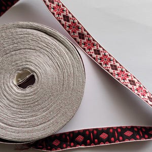 Könnte beinhalten: Eine Rolle graues Band mit dekorativem Rand. Das Band hat ein sich wiederholendes Muster aus roten und schwarzen geometrischen Formen. Das Band ist abgerollt und zeigt ein detailliertes Design. Das Band wird wahrscheinlich für Bastel- oder Nähprojekte verwendet.