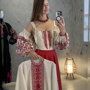 Può includere: Una camicia di lino bianca con ricami rossi e neri. La camicia ha un design tradizionale ucraino. La camicia è su un appendiabiti ed è tenuta da una persona.