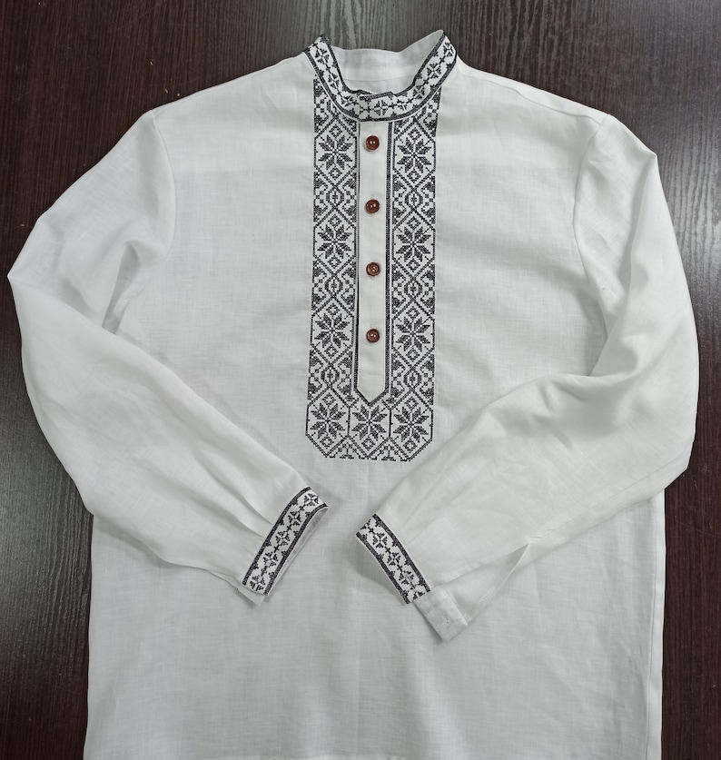 May include: White long-sleeved shirt with intricate black embroidery around the collar, down the front, and on the cuffs. The shirt features a mandarin collar and is fastened with brown buttons. The embroidery has a geometric floral pattern.