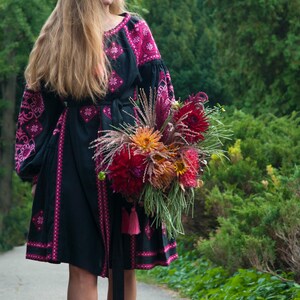Op de afbeelding: Een zwarte jurk tot op de knie met lange mouwen en felroze geometrische borduurpatronen. Het kledingstuk heeft een bijpassende ceintuur met roze kwastjes en een ronde halslijn. Een groot, kleurrijk boeket dahlia's wordt aan de voorzijde gehouden.